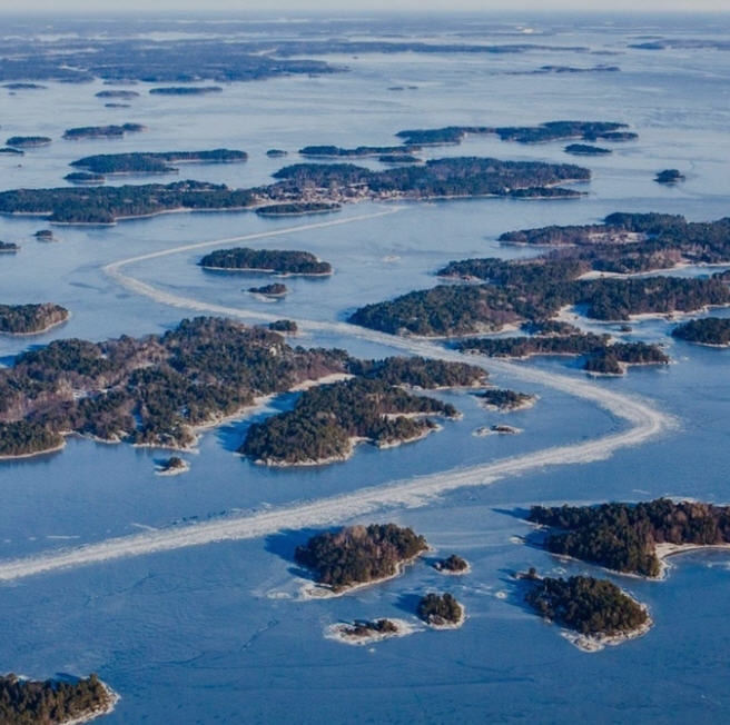 Rocky shoreline of a Stockholm Archipelago island, with pine trees, tranquil waters, and traditional red Swedish cabins.