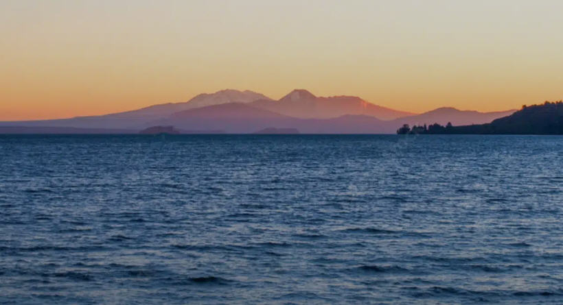 Shimmering expanse of Lake Taupō, New Zealand, surrounded by lush hills and snow-capped mountains.