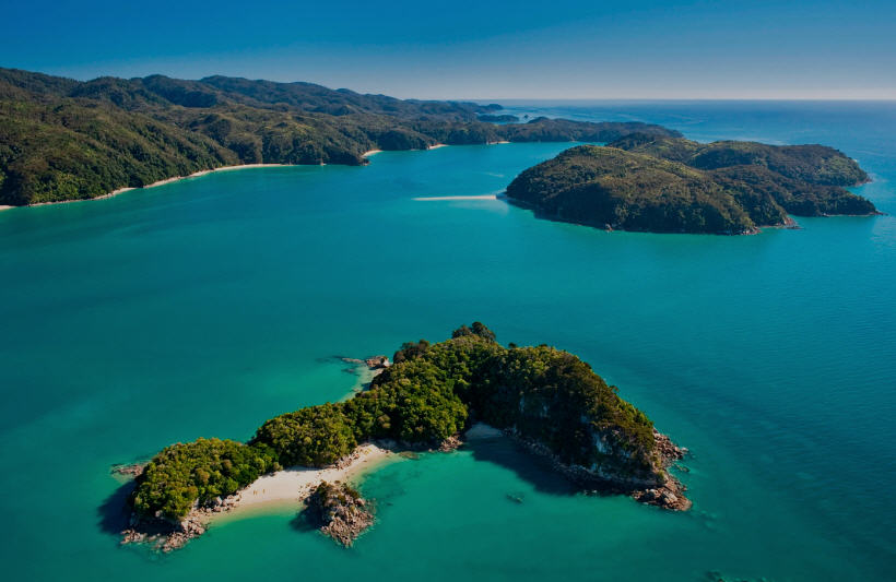 Panoramic view of a sunset over Abel Tasman National Park, casting long shadows on the coastline and turning the sky vivid shades of orange and pink.