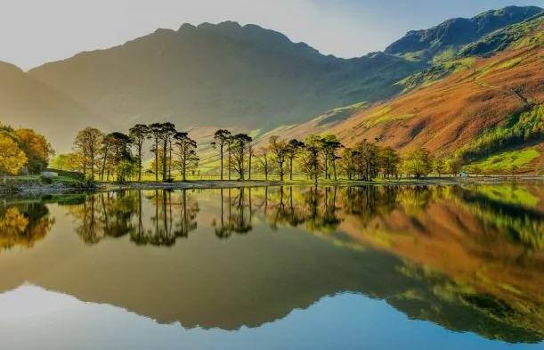Kayaker paddling across a calm Lake District lake at sunrise, surrounded by mist-covered hills.