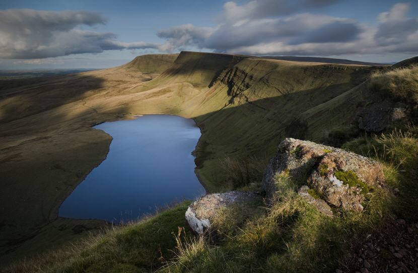 Panoramic view of the Brecon Beacons, showcasing rugged peaks, heather-covered slopes, and a reservoir nestled in a valley.